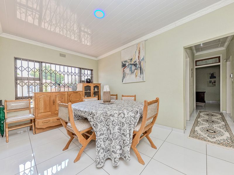 A dining room featuring white tiled flooring, coved ceiling details, and a security gate on the window, leading into a hallway.