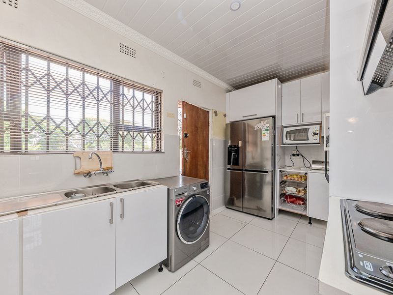 A functional kitchen featuring white cabinetry, tiled flooring, a stainless steel sink, and integrated appliances, with a security gate on the window.