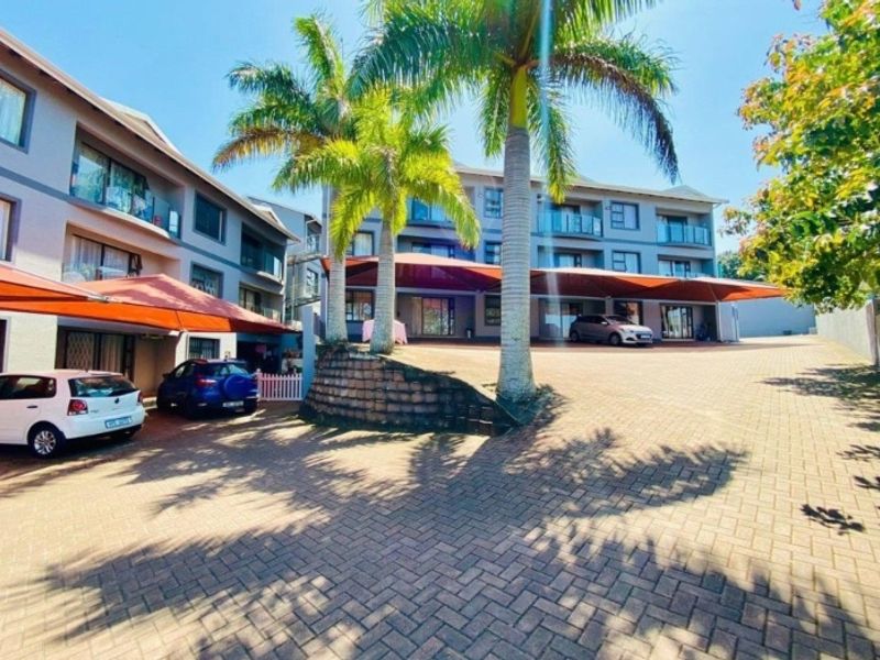 Exterior view of an apartment complex featuring a paved driveway, covered parking areas with shade sails, and multi-story residential units with balconies.