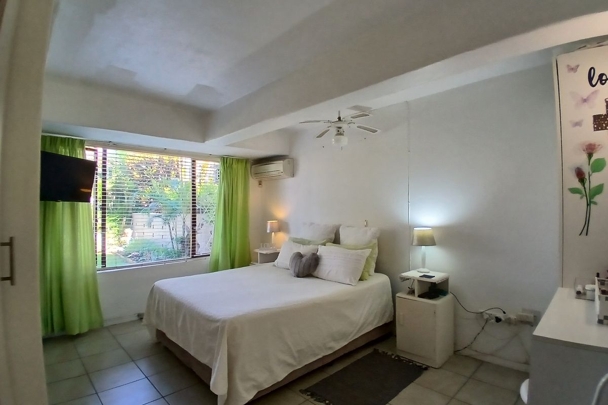 A bedroom featuring tiled flooring, a ceiling fan, and a wall-mounted air conditioning unit, with large windows overlooking a garden area.