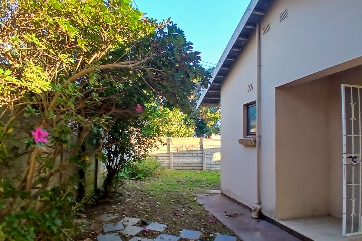 Rear exterior view of the property featuring a paved walkway, security gate, and a concrete block boundary wall with surrounding garden area.