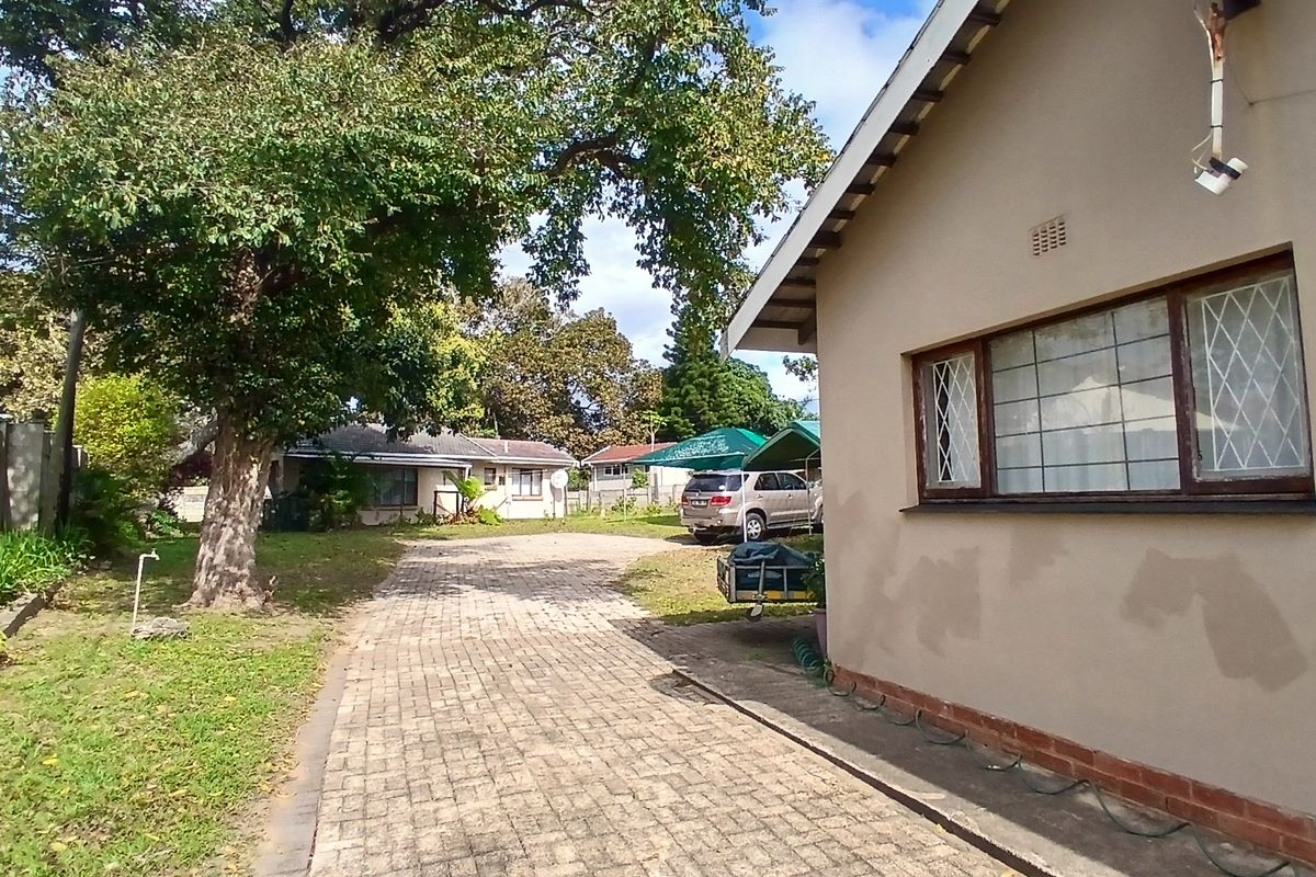 Rear exterior view of the property featuring a paved driveway, mature trees, and a side facade with security bars on the windows.