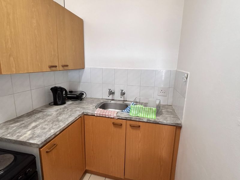 A compact kitchen featuring wooden cabinetry, laminate countertops, a stainless steel sink, and tiled flooring with a tiled backsplash.