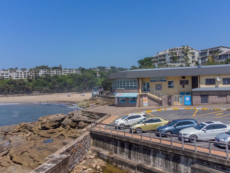Exterior view of a coastal property featuring a paved parking area, retaining wall, and direct access to the beach and oceanfront.