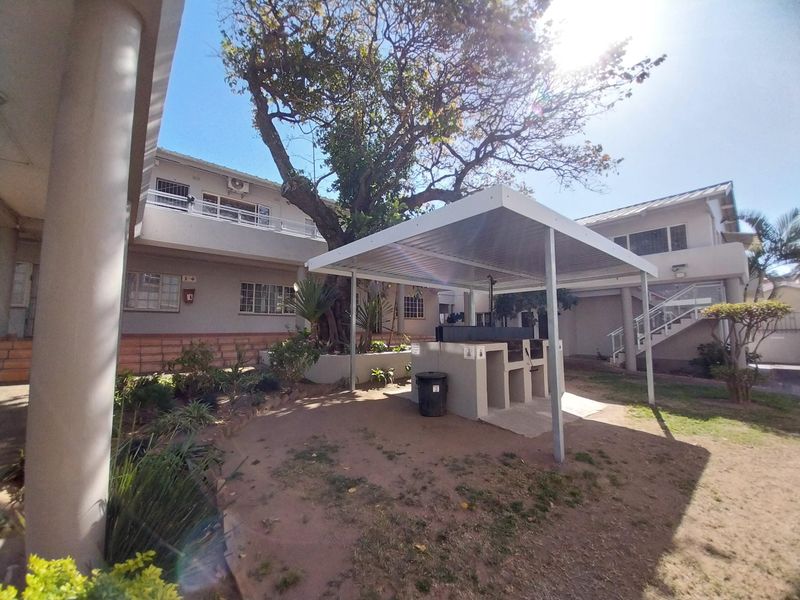 Communal outdoor area featuring a covered built-in braai station, paved ground, and established trees within an apartment complex setting.