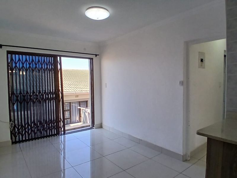 An empty open-plan living area featuring tiled floors, a sliding glass door with a security gate, and access to an adjacent kitchen space.