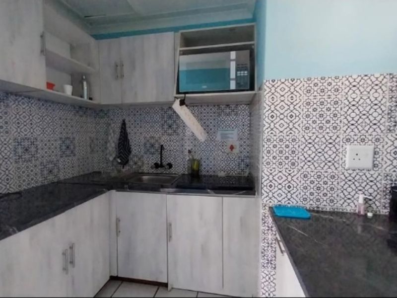 Compact kitchen featuring light wood-look cabinetry, dark countertops, and a decorative patterned tile backsplash with integrated microwave and sink.
