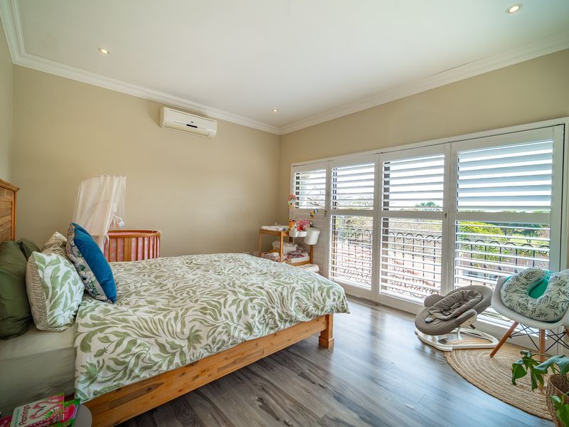A bright and well-maintained bedroom featuring laminate flooring, recessed lighting, a split-system air conditioner, and plantation shutters opening onto a balcony.