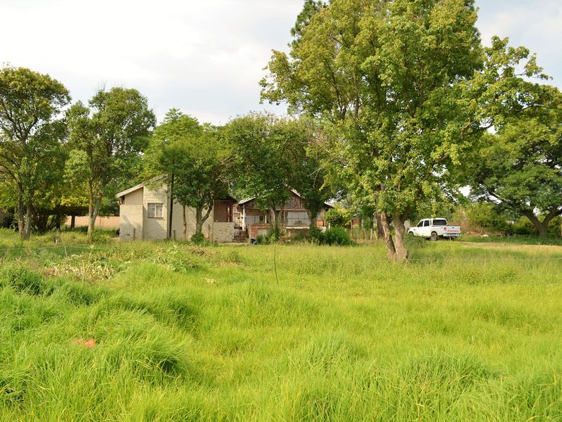 A view of a residential property set within a large, grassy plot featuring mature trees and an existing dwelling.