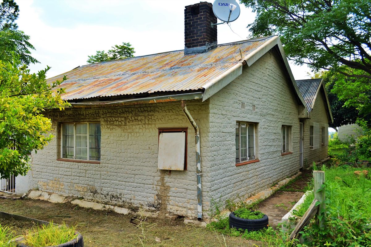 Rear view of a residential structure featuring a corrugated metal roof, brick chimney, and textured masonry walls showing signs of wear and weathering.