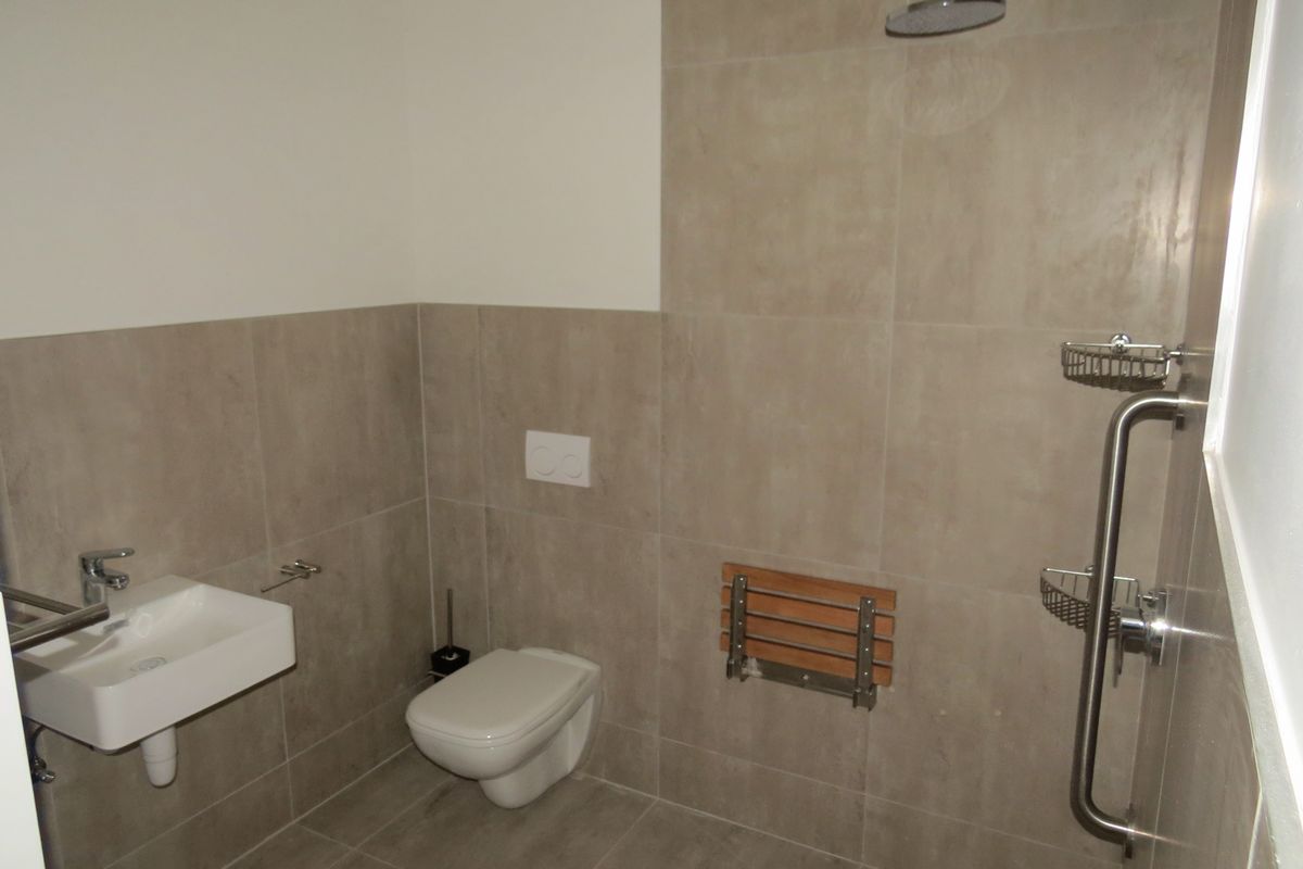 A modern, accessible bathroom featuring neutral wall tiles, a wall-mounted basin, a wall-hung toilet, and a walk-in shower with grab rails and a foldable seat.
