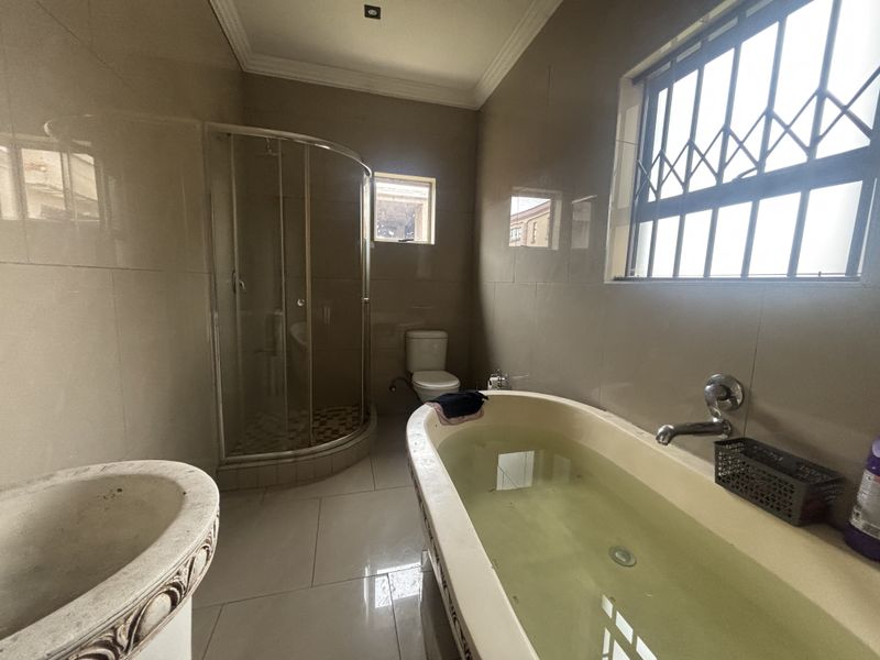 A functional bathroom featuring a curved glass shower, a freestanding bathtub, a pedestal basin, and tiled finishes throughout with security bars on the window.