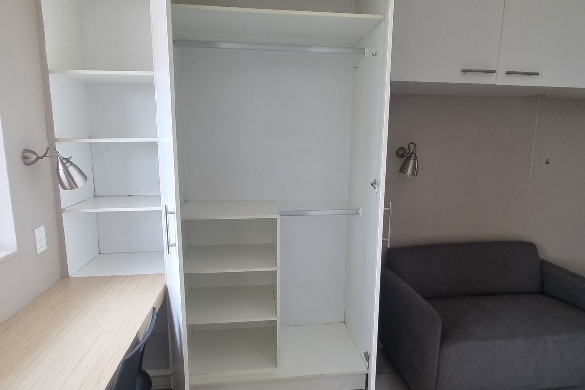 A bedroom featuring a built-in desk, integrated shelving, a wardrobe, and overhead storage cabinets with wall-mounted lighting.