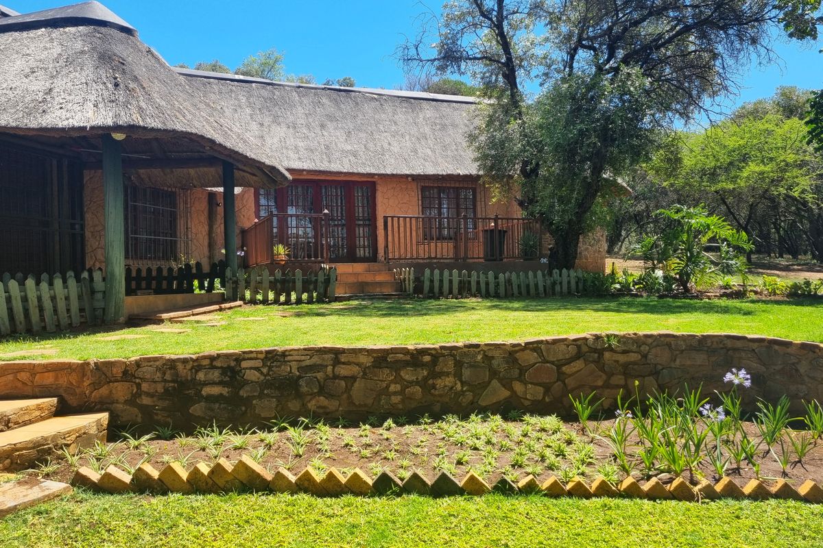 A charming rear exterior view featuring a traditional thatch roof, a raised timber deck, and a well-maintained stone retaining wall within a landscaped garden.