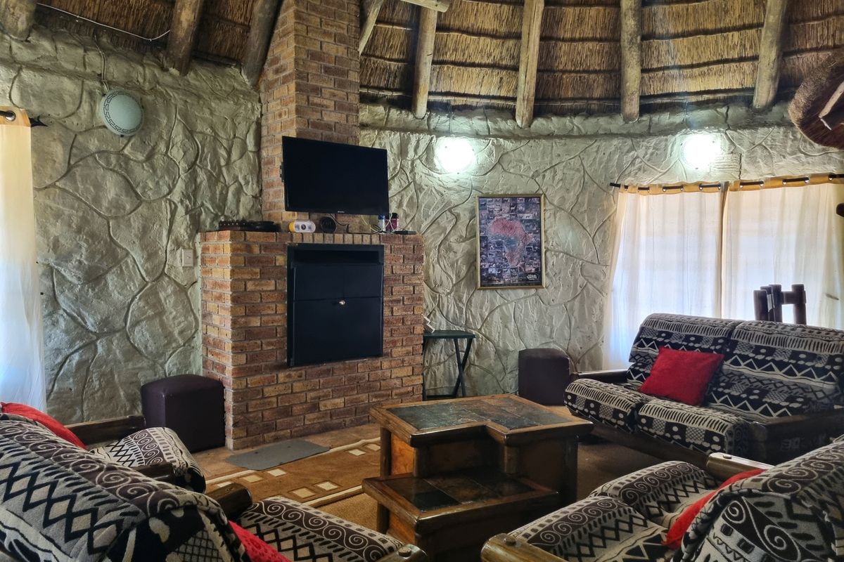 A rustic living room featuring a central brick fireplace, distinctive textured stone walls, and a traditional thatched roof with exposed timber rafters.