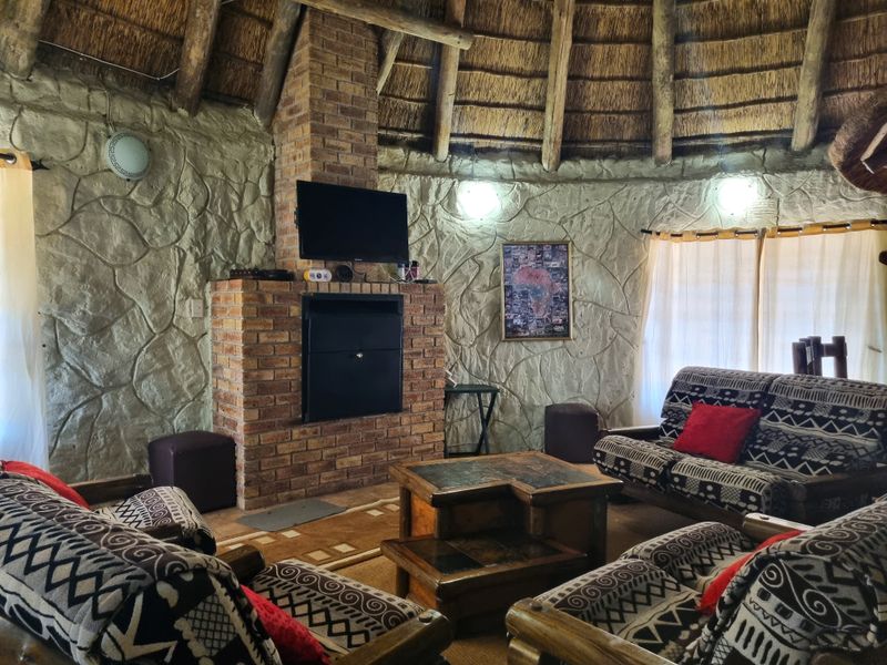 A rustic living room featuring a central brick fireplace, distinctive textured stone walls, and a traditional thatched roof with exposed timber rafters.