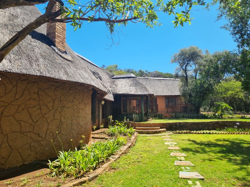 A view of the rear exterior featuring a traditional thatched roof, textured facade, stone retaining walls, and a landscaped garden with a stone pathway.