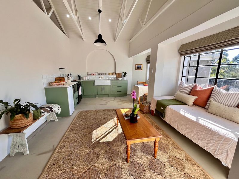 Bright open-plan living and kitchen area featuring exposed vaulted ceilings, farmhouse sink, sage green cabinetry, and large steel-framed windows allowing ample natural light.