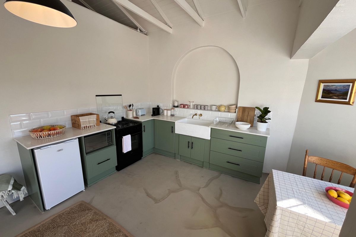 A charming cottage-style kitchen featuring sage green cabinetry, a farmhouse sink, subway tile backsplash, and exposed white rafters with an arched wall feature.