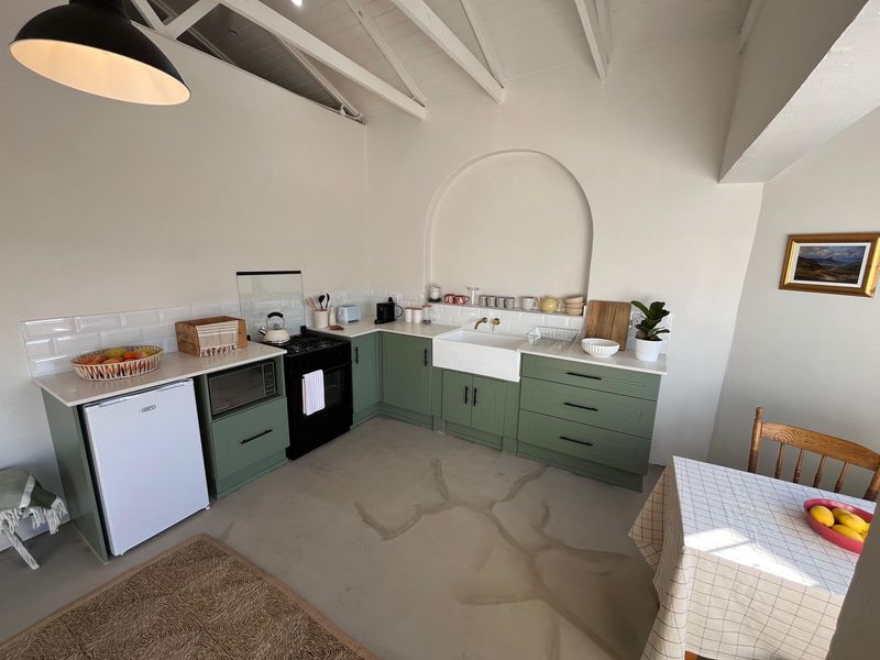 A charming cottage-style kitchen featuring sage green cabinetry, a farmhouse sink, subway tile backsplash, and exposed white rafters with an arched wall feature.