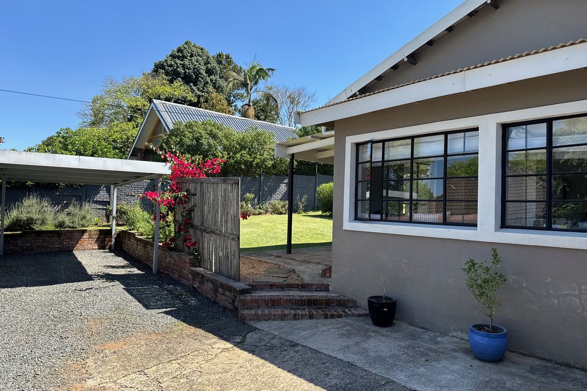 Rear exterior view of the cottage featuring a carport, gravel driveway, brick retaining walls, and steel-framed windows overlooking a garden area.