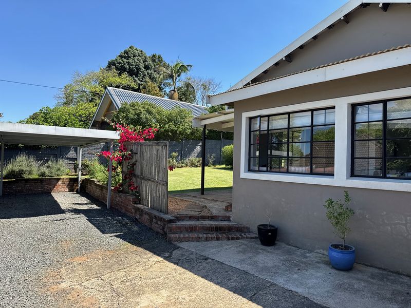 Rear exterior view of the cottage featuring a carport, gravel driveway, brick retaining walls, and steel-framed windows overlooking a garden area.