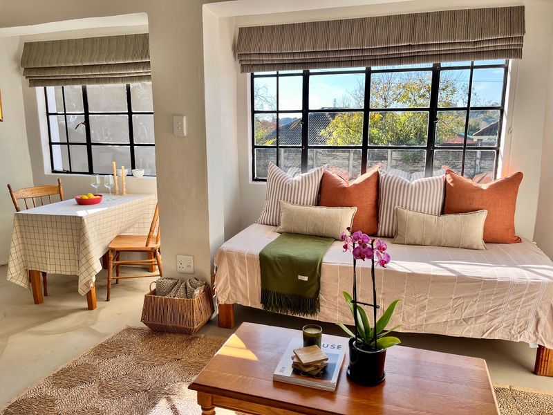 Bright open-plan living and dining area featuring large steel-framed windows, neutral concrete flooring, and classic roman blinds.