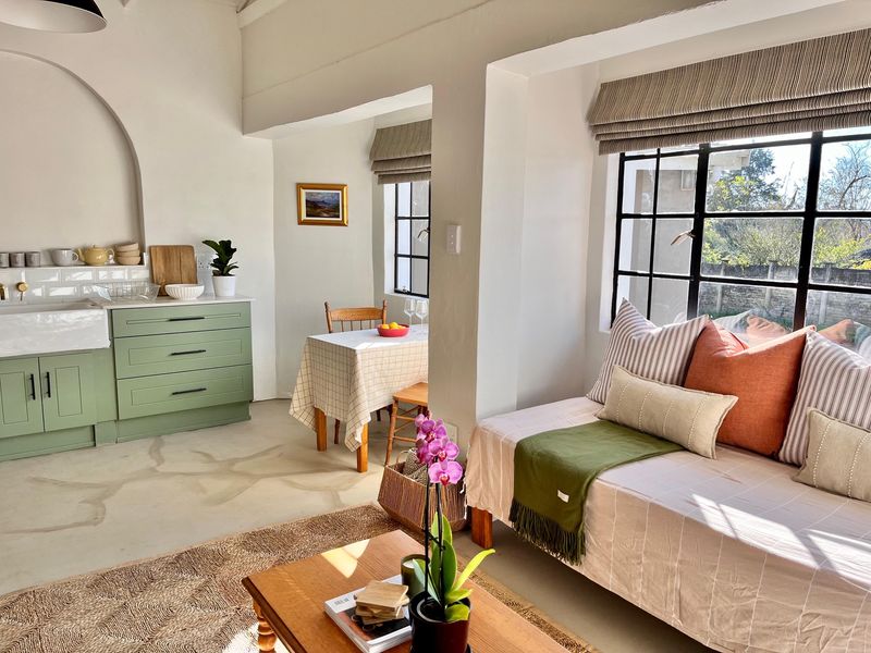 Bright open-plan living and kitchen area featuring sage green cabinetry, a farmhouse sink, concrete floors, and large steel-framed windows with natural light.