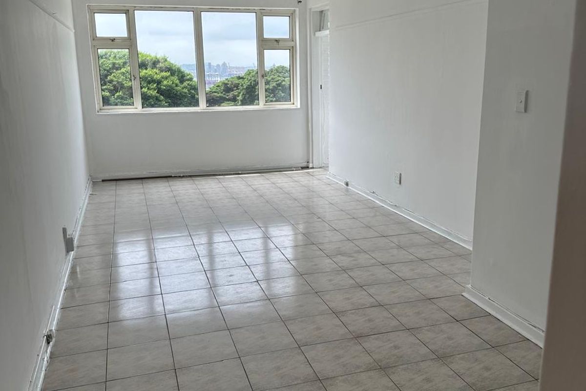 An empty, sunlit living area featuring tiled flooring and a large window with a view, offering a blank canvas for renovation.