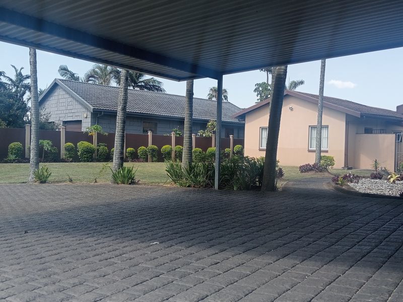 A view from under a covered carport looking out onto a paved driveway, landscaped garden, and neighboring cottage-style residential properties.