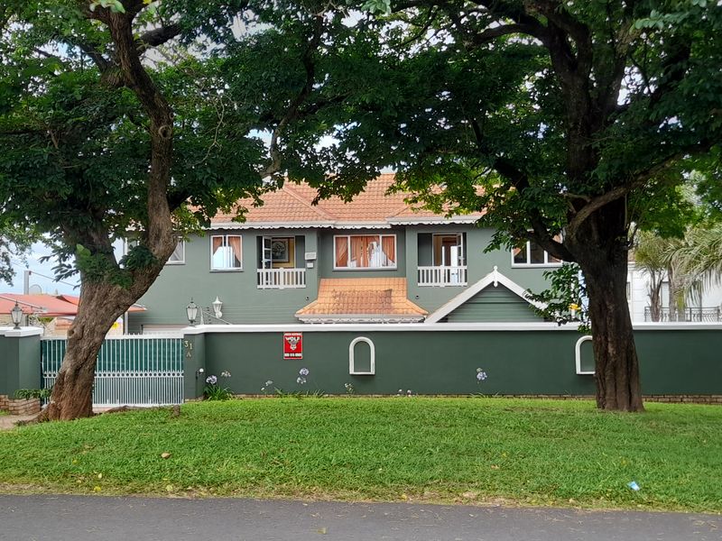 Front elevation of a two-story house featuring a green facade, terracotta roof tiles, and a secure perimeter wall with gated access.