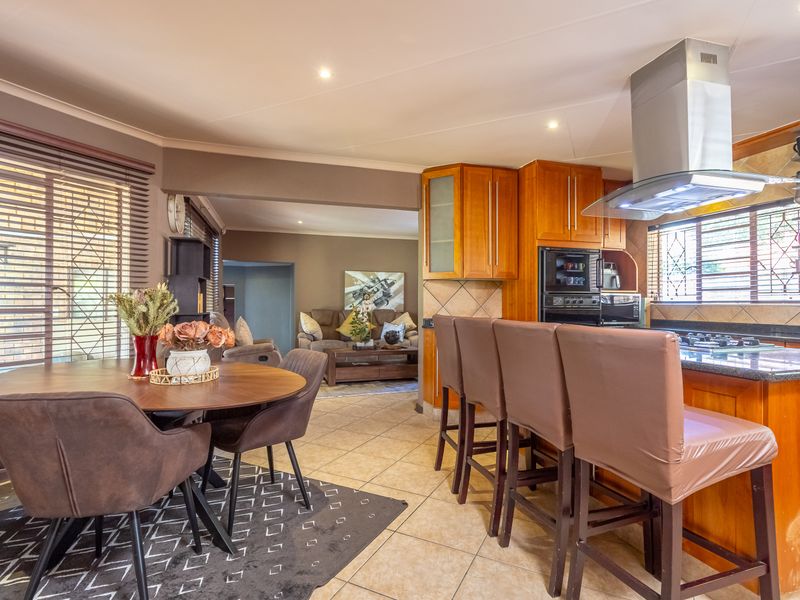 Open-plan kitchen and dining area featuring wooden cabinetry, granite countertops, built-in appliances, and tiled flooring, flowing into an adjacent living space.