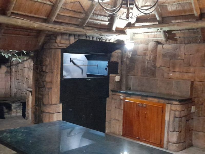 Covered outdoor entertainment area featuring a built-in braai, stone-clad walls, thatch roof, and a granite-topped prep counter with wooden cabinetry.