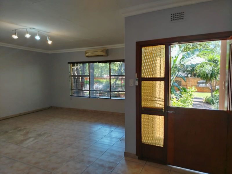 Spacious living area featuring tiled flooring, a wall-mounted air conditioning unit, and a wooden door with security mesh leading to the garden.