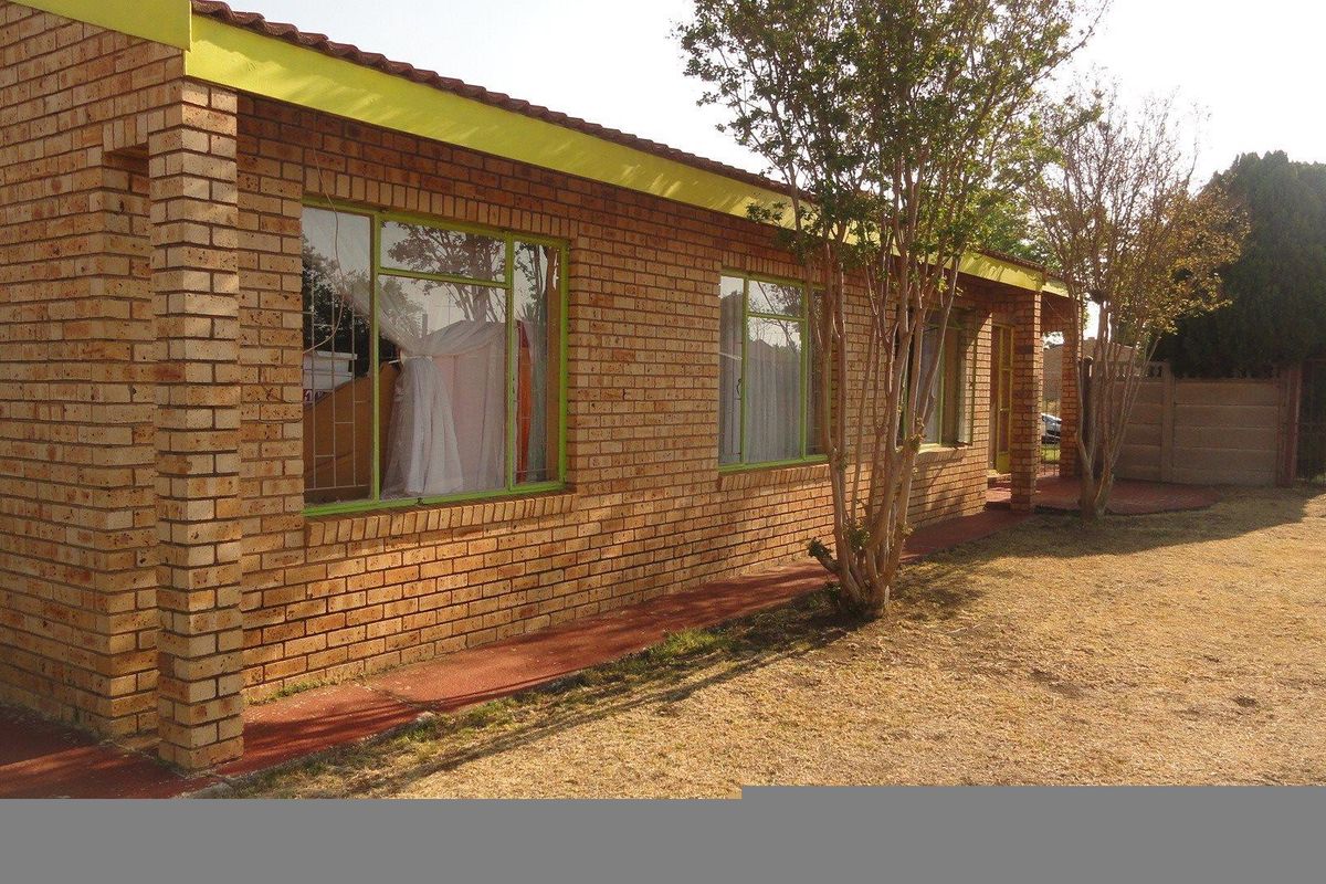 A side view of a single-story face-brick house featuring large windows with green frames and a paved walkway along the exterior.