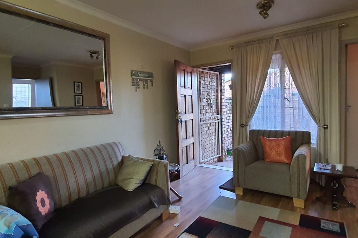 A living room featuring laminate flooring, a wooden entrance door with security gate, and access to an adjacent room.