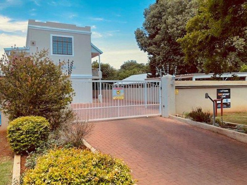 Exterior view of the apartment complex entrance featuring a paved driveway, secure white metal gate, and perimeter wall with security fencing.