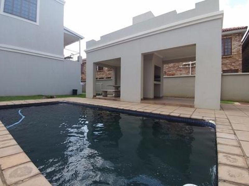 Outdoor pool area featuring a covered patio with a built-in braai, paved surround, and landscaped garden space.
