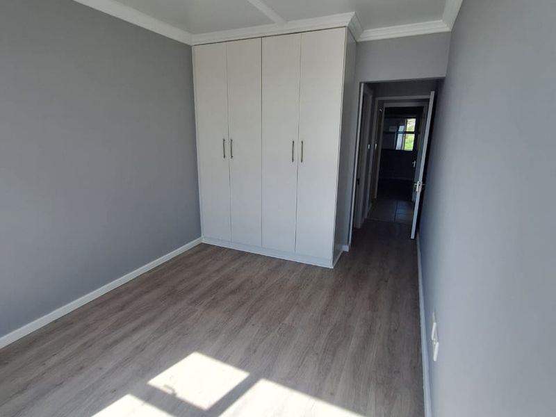 A bright, empty bedroom featuring modern laminate flooring, neutral grey walls, and built-in white cupboards, offering a clean and versatile living space.
