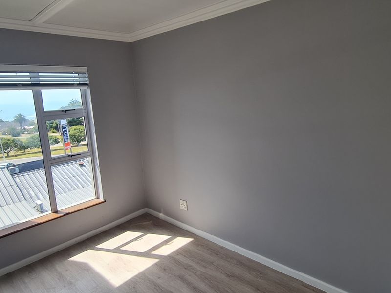 A bright, empty bedroom featuring modern laminate flooring, built-in cupboards, and a window with blinds offering a pleasant exterior view.