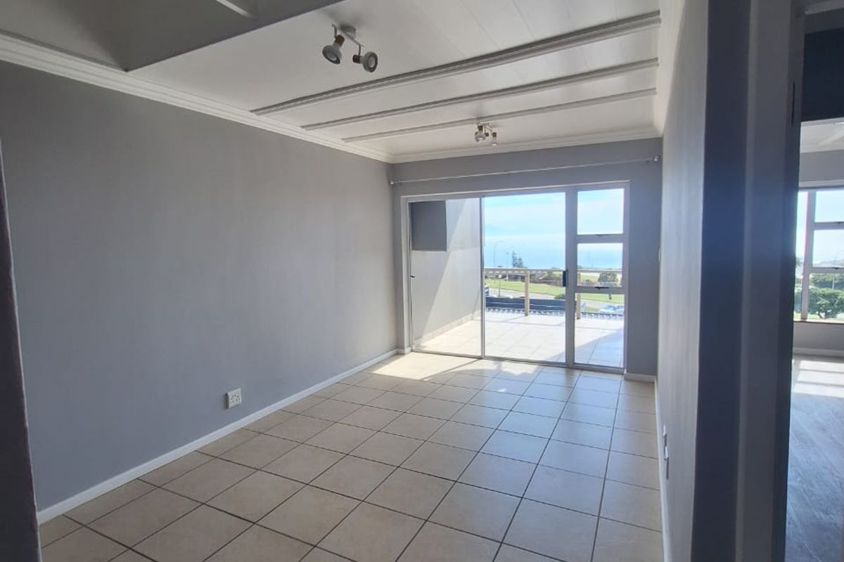 A bright, empty living room featuring neutral tiled flooring, exposed beam ceilings, and large sliding glass doors leading to an outdoor balcony with views.
