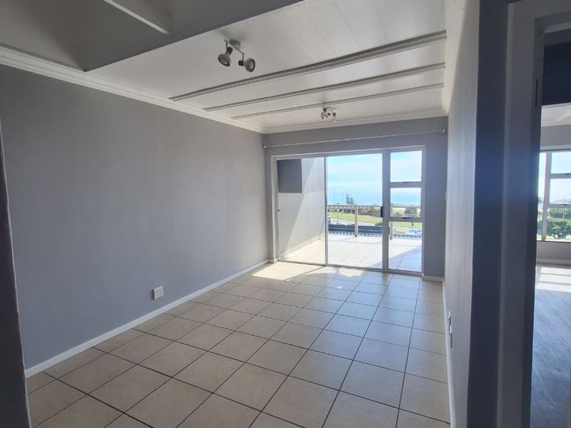 A bright, empty living room featuring neutral tiled flooring, exposed beam ceilings, and large sliding glass doors leading to an outdoor balcony with views.