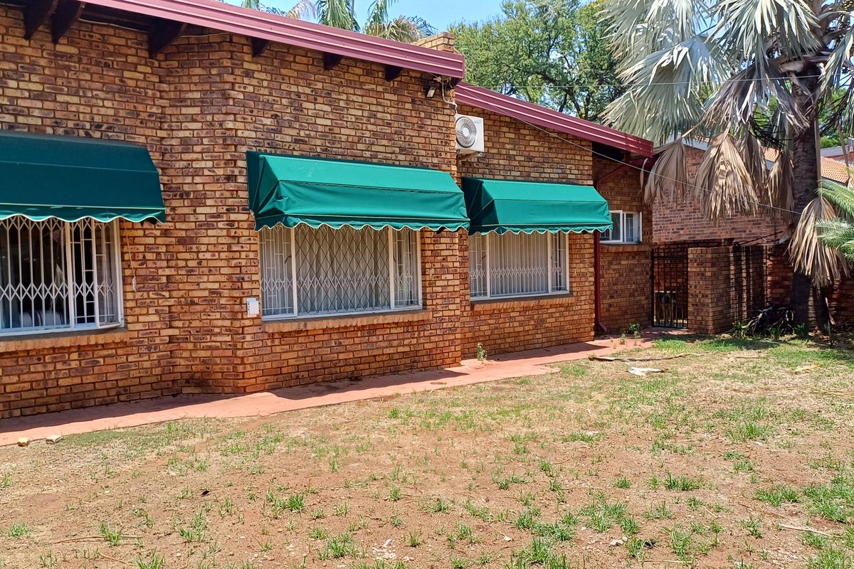 Rear view of a face brick house featuring window awnings, security gates, and an outdoor paved area with a garden space.