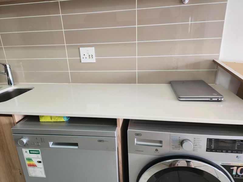 A functional laundry area featuring a stone countertop, tiled backsplash, stainless steel sink, and space for integrated appliances.