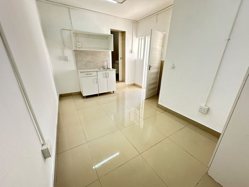 A compact, empty kitchenette area featuring tiled flooring, basic cabinetry with a sink, and exposed electrical conduit, providing a functional utility space.