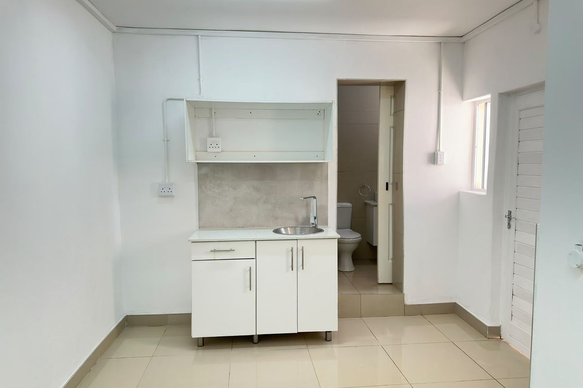 A compact kitchenette featuring white cabinetry, a stainless steel sink, and tiled flooring, with direct access to an adjoining bathroom.