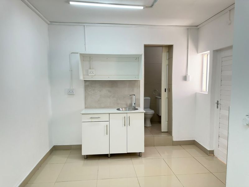 A compact kitchenette featuring white cabinetry, a stainless steel sink, and tiled flooring, with direct access to an adjoining bathroom.
