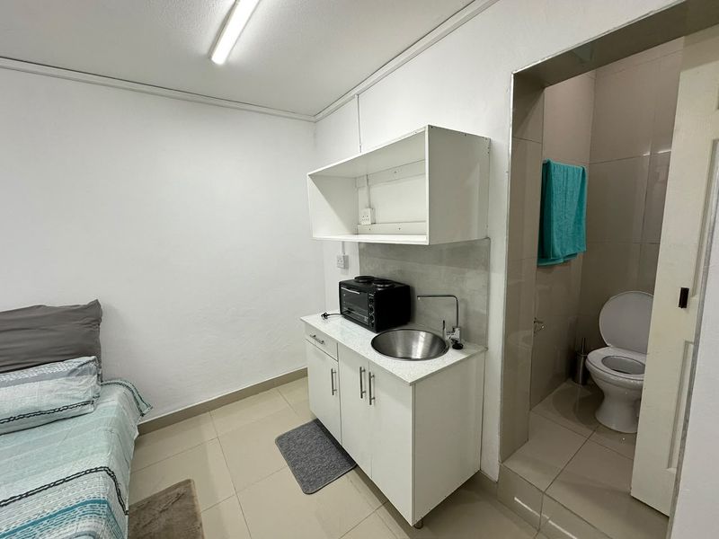 Compact kitchenette area featuring white cabinetry, a stainless steel sink, and direct access to an adjoining bathroom with tiled finishes.