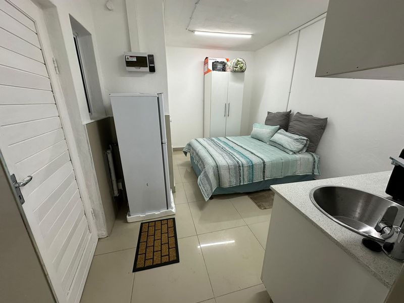 Compact open-plan studio space featuring tiled flooring, a small kitchenette area with a stainless steel sink, and direct access via a louvered door.