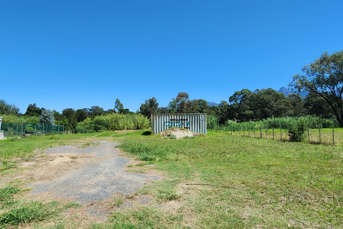 A view of a vacant residential plot featuring a shipping container, perimeter fencing, and established trees in a rural setting.